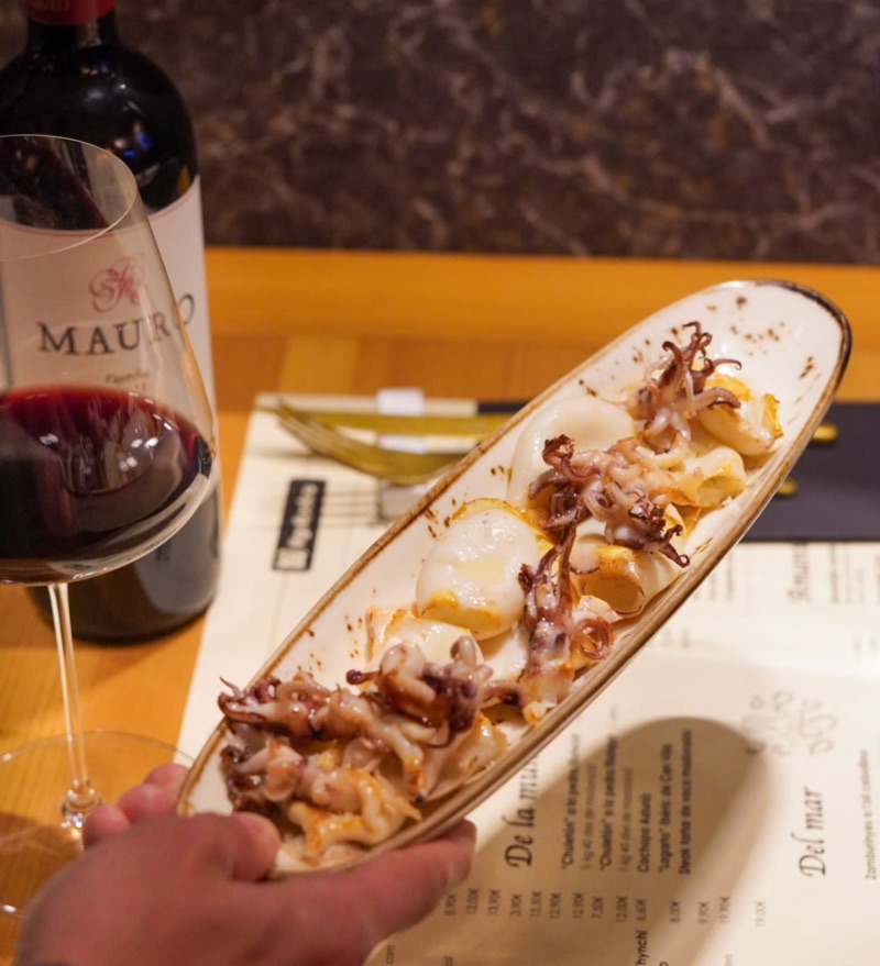 Una mà acosta una safata de calamars, amb una ampolla i una copa de vi negre