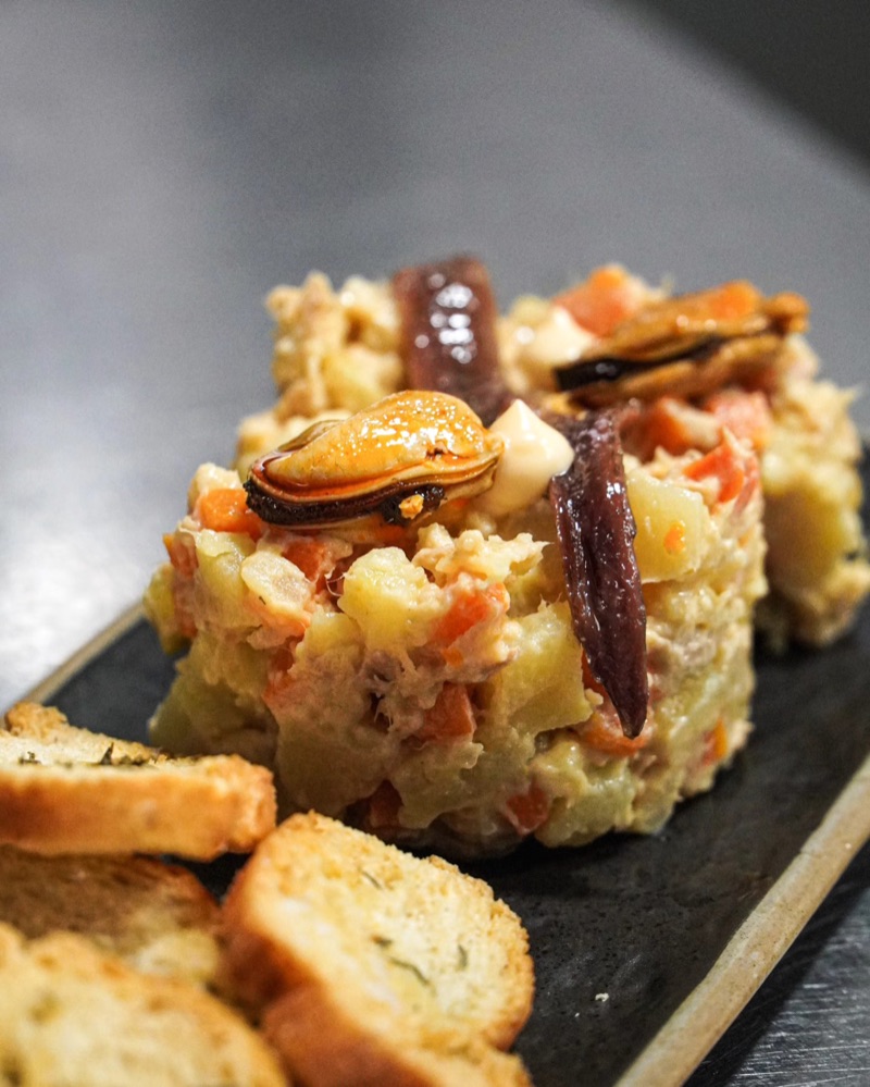 Ensaladilla amb musclos i anxoves, amb torrades, sobre una pissarra