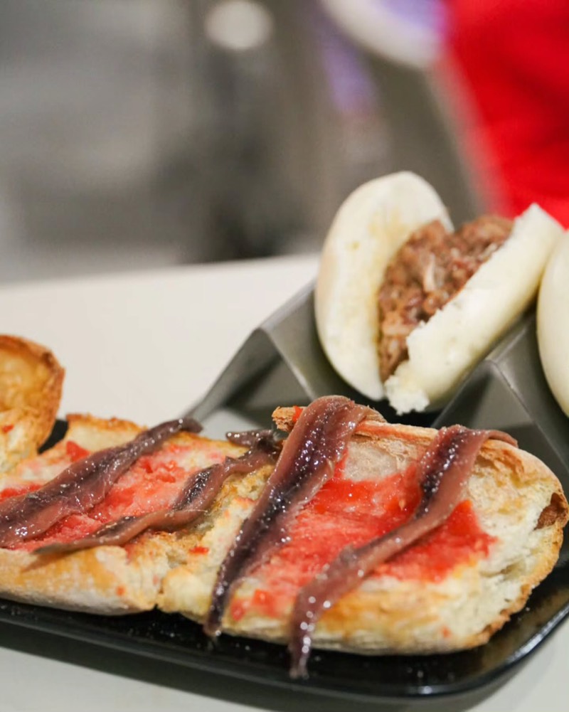 Coca amb tomàquet i anxoves, amb un pa bao al darrere